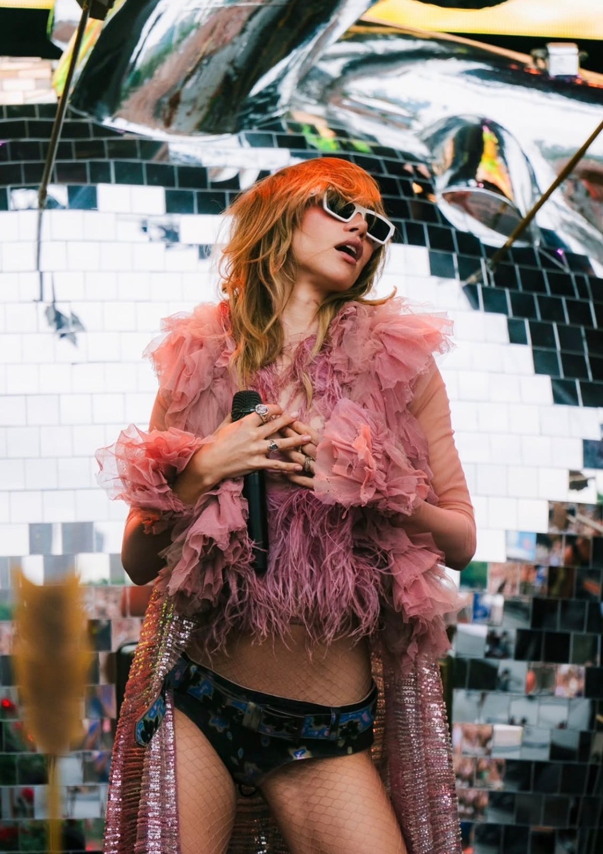 A woman with bright orange hair and white sunglasses stands in front of a mirrored tile wall, holding a microphone. She is wearing a voluminous pink feathered top and a shimmering pink open-front cardigan over dark patterned bikini bottoms.
